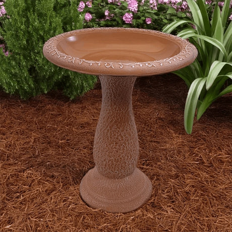 Terra cotta pedestal birdbath in mulch bed surrounded by flowers and greenery