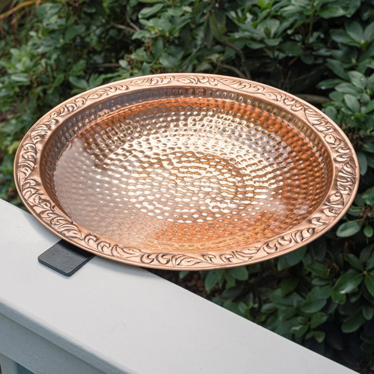 A hammered copper bird bath tray with an ornate leaf-patterned rim, shown resting on a white railing with green foliage in the background.